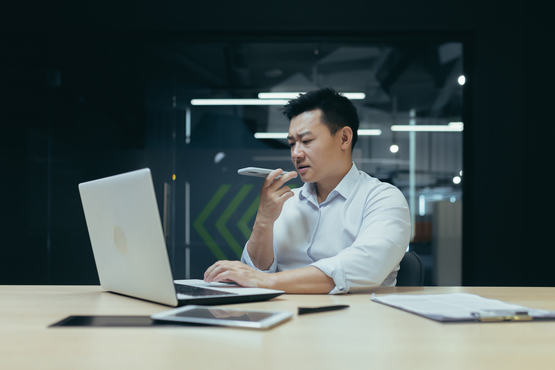 A young Asian man is sitting in the office at the table, working on a laptop. Talking on the phone through the loudspeaker. records a voice message.