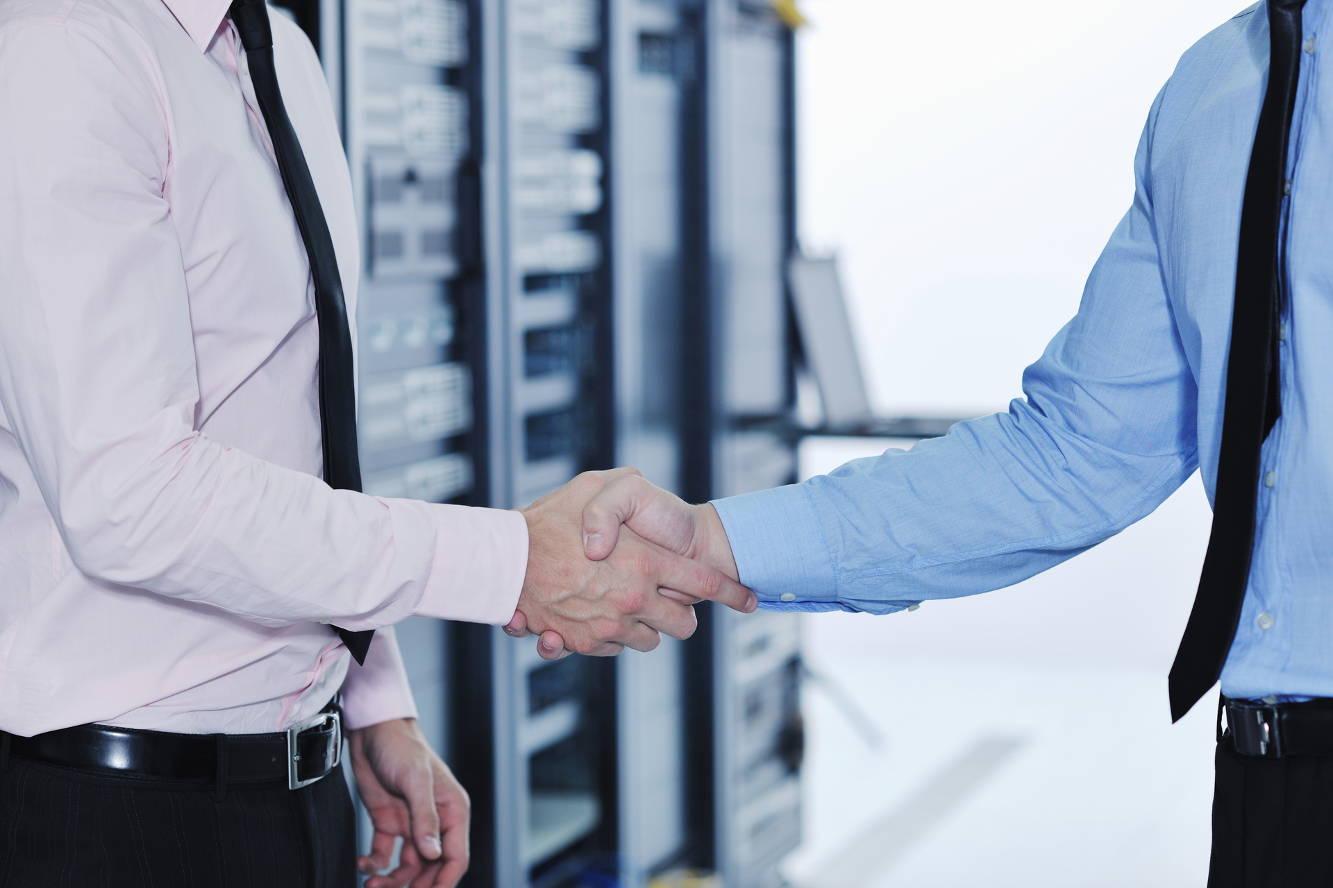 Two businesspeople in dress shirts and ties shaking hands in a modern office setting with large windows in the background.Retry